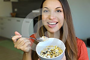 Close up of beautiful young woman eating skyr yogurt with cereal muesli fruit at home, looking at camera, focus on the model eyes