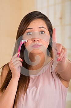 Close up of a beautiful young woman holding a shaver with onde hand and pointing to the sky with her other hand, in a