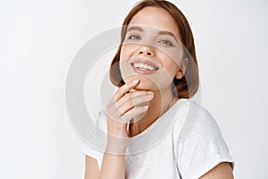 Close up of beautiful young woman with natural light make-up fresh face, touching chin and smiling tenderly, standing