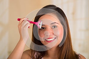 Close up of a beautiful young woman using a shaver on her forehead, in bath background