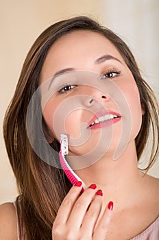 Close up of a beautiful young woman using a shaver to shave her cheek, in a bath background