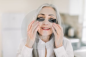 Close up beauty portrait of smiling pretty senior gray haired woman, applying under eye mask, black patches on her face