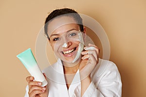 Young woman with beautiful toothy smile applying a facial cream or scrub on her face standing isolated over beige background with