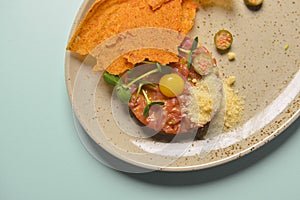 Close up of beef tartar with capers and small toasts. Served on a gray textured plate over pastel blue mint background.