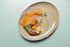 Close up of beef tartar with capers and small toasts. Served on a gray textured plate over pastel blue mint background.