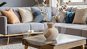 Close up of beige ceramic vase with twigs on rustic live edge accent coffee table against grey sofa with fabric pillows.