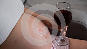 Close-up of the belly of a pregnant woman holding a glass of red wine while sitting on the sofa. Skin rash.