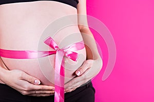 Close-up belly of pregnant woman with satin ribbon tied around it in the form of bow as gift on pink background