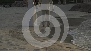 Close up below of the body, Bride and Groom are holding and walking on the beach beside the sea and wave in the Twilight time