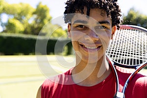 Close-up of biracial young man with tennis racket smiling in tennis court and looking at camera photo