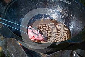 Close-up, in a black cast-iron cauldron, vegetable oil boils on fire and raw pieces of pork ribs are put into it with tongs. Food