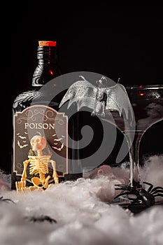 Close-up of black Halloween cocktail glass with bat, skeleton and bottle of poison on spider web, black background,