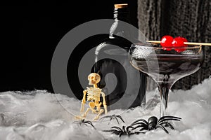 close-up of black Halloween cocktail glass with red cherries, skeletons, spiders and dark bottle on spider web, black background,