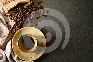 Close-up of Black hot coffee with milk foam in black ceramic cup and napkin with coffee beans roasted in a sack on dark stone tabl