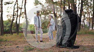 Close-up black rucksack in forest with blurred happy couple talking standing at background. Unrecognizable travelers