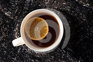 Close-up black tea with lemon slice in white cup on background from knitted sweater, horizontal. Hot drink