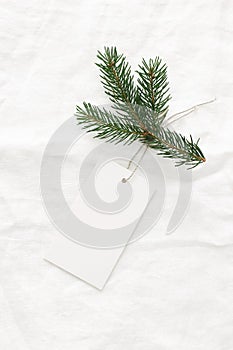 Close-up of blank white paper gift tag with rope and green fir branch on white linen tablecloth background. Price tag photo