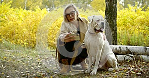 Close - up of a blond young girl in a coat and boots. She is in the woods, squatting next to his big white dog and makes