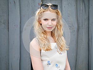 Close up of blonde student girl with trendy sunglasses posing against grey wooden , enjoying warm sun light, recreation and relaxa
