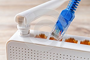 Close-up of blue network cable plug almost inserting into the socket of the white Wi-Fi wireless router on a wooden desk. Home and