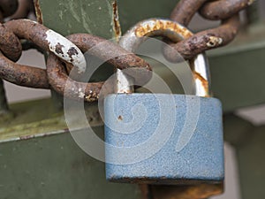 Close up of blue padlock with rusty chain locked at old green gate door, selective focus