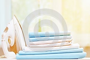 Close-up of blue and white clean bedding on a blurred background. A stack of folded bedsheets on the table, an iron stands nearby.