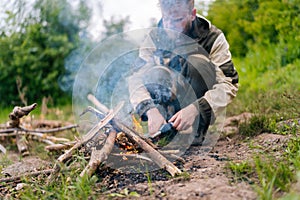 Close-up of blurred survivalist male in raincoat starting fire using gas lighter on bank of river for cooking and photo