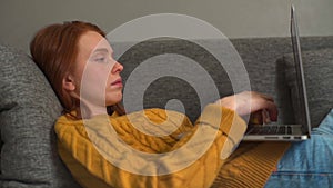 Close-up of bored Caucasian young woman typing on laptop computer while lying on couch sofa at home.