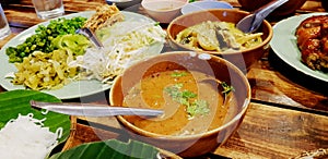 Close up bowl of spicy fish curry sauce on wooden table with Thai rice noodle, vegetable, grilled chicken and green curry