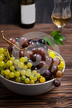 Close up bowl of various grapes: red, white and black berries on the dark wooden table with bottle and glass of white wine in the