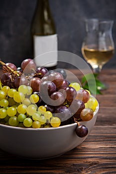 Close up bowl of various grapes: red, white and black berries on the dark wooden table with bottle and glass of white wine in the