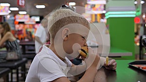 Close-up of a boy eating a burger sitting at a table in a shopping center.