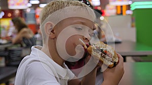 Close-up of a boy eating a burger sitting at a table in a shopping center.