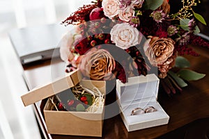 Close up of bridal bouquet of pink, orange and red flowers, butonniere and two golden wedding rings in box on table indoors.