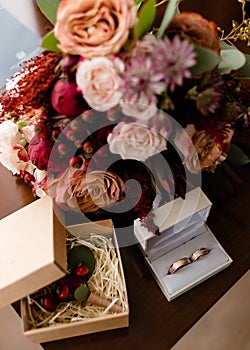 Close up of bridal bouquet of pink, orange and red flowers, butonniere and two golden wedding rings in box on table indoors.