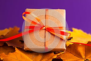 Close up of brown gift box with a orange satin ribbon bow on the yellow leaves on vibrant purple blurred background with copy
