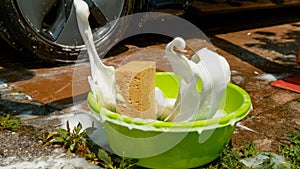 CLOSE UP: Brown sponge falls into a bucket full of soapy water next to a car.