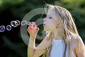 Close-up of bubble-blowing young girl 11, with long blond hair, blowing brightly colored bubbles in the garden.