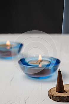Close-up of burning incense on wood, candles with blue glass, selective focus, on white table, black background