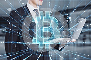 Close up of businessman hand using laptop with abstract glowing holographic bitcoin icon on blurry office interior background.