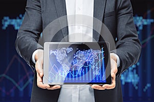 Close up of businessman hands holding tablet with map and glowing upward candlestick forex chart on dark grid background. Trade,