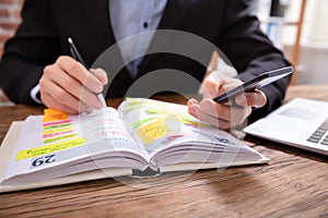 Businessman Holding Cellphone Writing Schedule In Diary With Pen