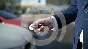 Close-up of businessman pressing car key. Action. Businessman opens car with electronic key and looks at phone. Modern