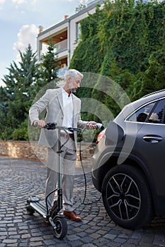 Close up of businessman in suit on way to work standing at eletric scooter and charging his electric car. Concept of eco