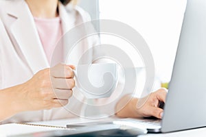 Close up of businesswoman using laptop computer and sitting at office desk, holding a cup of coffee