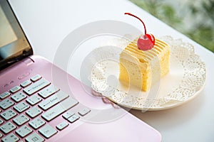 Close up butter chiffon cake with cherry fruit on top in a white plate next to a pink laptop on a white desktop next to the window