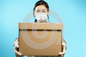 Close-up of a cardboard box in the hands of a young woman in a medical protective mask and gloves on a blue background