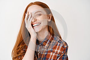 Close-up of carefree female model with ginger hair and blue eyes, covering half of face and laughing with happy face at