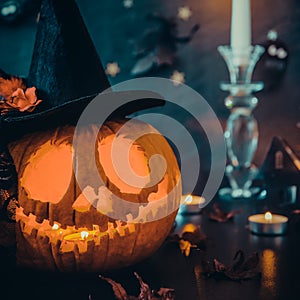 Close up carved pumpkin in witch hat, burning candles and paper silhouettes of bats, castle, ghosts on black stone background.