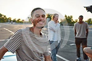 A close up of a casually dressed dark-skinned latina guy with a piercing smiling looking into a camera standing outside